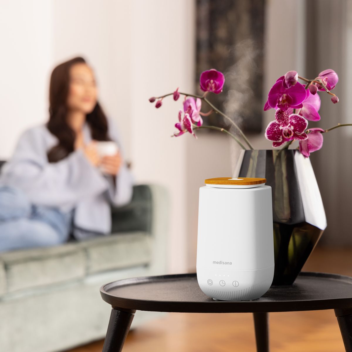 Ein weißer Aroma-Diffuser steht auf einem Tisch neben einer Vase mit Orchideen, während im Hintergrund eine Frau entspannt sitzt.